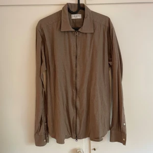 Beige overshirt från John Henric - Stilren beige overshirt från John Henric med dragkedja framtill och klassisk krage. Skjortan har långa ärmar med manschettknappar och är tillverkad i ett mjukt bomullstyg. Perfekt för en clean och avslappnad look. Nypris 899kr