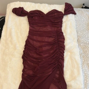 Vinröd långklänning med offshoulder - Säljer en vinröd långklänning med snygg offshoulder-design och draperad mesh över hela klänningen. Klänningen har hjärtformad urringning, markerad midja och slits framtill. Perfekt för dig som vill sticka ut med en elegant och trendig look.