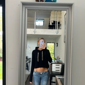 Svart croppad hoodie med glittriga snören - Svart croppad hoodie med långa ärmar och huva. Tröjan har breda glittriga snören framtill som ger en extra touch. Ribbad nederkant och ärmslut. Perfekt för dig som gillar en chill och trendig stil.