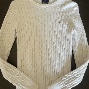 Vit kabelstickad tröja från Gant - Vit kabelstickad tröja från Gant i stretchig bomull. Klassisk rund halsringning och diskret Gant-logo broderad på bröstet. Långärmad modell med ribbade muddar och en clean, tidlös look. 
