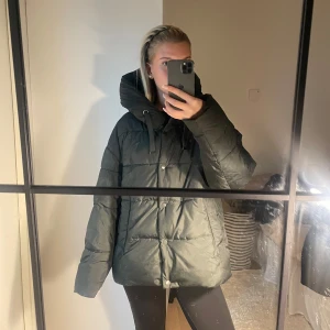 Mörkgrön pufferjacka med hög krage - Säljer en mörkgrön pufferjacka med hög krage och snörning. Jackan har en oversized passform, långa ärmar och knappar framtill. Perfekt för kalla dagar med sin quiltade design och stora fickor. Snygg 