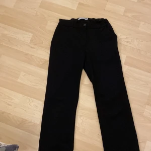 Svarta kostymbyxor från 157 - Svarta kostymbyxor från 157 med klassisk rak passform och knappstängning i midjan. Byxorna har bälteshällor och är tillverkade i ett mjukt tyg som känns bekvämt hela dagen. Perfekta för en clean och stilren look.