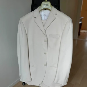 Simbols Culture kostym – Ljusbeige, strl 48 - Stilren ljusbeige kostym från Simbols Culture. Tillverkad i 100% bomull vilket gör den lätt, luftig och perfekt för vår och sommar.  Klassisk design med treknäppt kavaj, bröstficka, två framfickor samt fyra dekorativa knappar vid ärmsluten. Matchande byxa med bakficka och bälteshällor. En tidlös kostym som passar utmärkt till bröllop, fest och högtidliga tillfällen.  Storlek: 48 Färg: Ljusbeige Material: 100% bomull Nypris: 3595 kr Skick: Helt ny med tags kvar