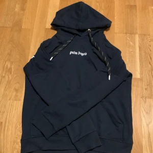 Mörkblå Palm Angels hoodie S - Säljer en mörkblå hoodie från Palm Angels i storlek S. Tröjan har stor vit logga på ryggen och mindre logga på bröstet. Klassisk huva med snörning och långa ärmar. Mjuk bomullskänsla och ribbade muddar för en clean streetstyle.