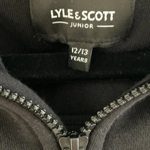 Svart hoodie från Lyle & Scott Junior - Svart hoodie från Lyle & Scott Junior med huva, halv dragkedja och känguruficka. Tröjan har en bröstficka med lock och klassisk gul logga på ärmen. Perfekt för dig som gillar en stilren och bekväm look.