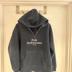Mörkblå fleecehoodie Peak Performance - Mysig mörkgrå fleecehoodie från Peak Performance med broderad logga på bröstet. Tröjan har huva, half zip och en stor magficka. Perfekt för kalla dagar och chill vibes.