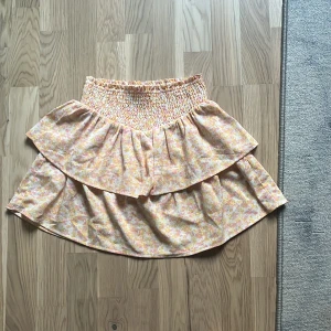 Blommig volangkjol från Kappahl - Supersöt kort volangkjol från Kappahl med blommigt mönster i ljusa toner av rosa, gul och vit. Kjolen har smock i midjan för extra bekväm passform och dubbla volanger som ger en flowy känsla. Knappt använd.