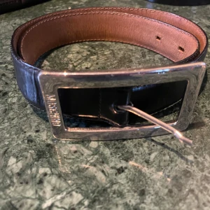 Svart skärp från Moschino i skinn - Snyggt svart bälte från Moschino i äkta skinn med en bred, rektangulär silverfärgad metallspänne där Moschino-loggan är ingraverad. Insidan är brun och har stilrena sömmar längs kanterna. Perfekt för att ge din outfit en lyxig touch.