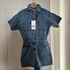Jeansshorts jumpsuit från Zara - Snygg jeansshorts jumpsuit från Zara i klassisk blå denim. Modellen har korta ärmar, krage, två stora bröstfickor och knappar framtill. Midjan är markerad och shortsen har både fram- och bakfickor. Den är helt oanvänd med lapp kvar!!🫶