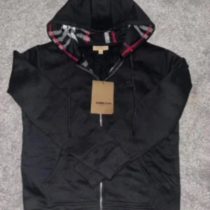 Svart Burberry hoodie med dragkedja - Svart hoodie från Burberry med klassiskt rutigt foder i huvan och broderad logga på bröstet. Tröjan har hel dragkedja, snörning i huvan och två fickor framtill. Perfekt för dig som gillar stilrena och exklusiva streetwear-plagg.