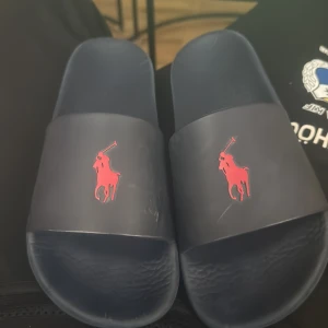 Mörkblå slides från Polo Ralph Lauren - Snygga mörkblå slides från Polo Ralph Lauren med röd logga framtill. Skorna har en bred rem över foten och är tillverkade i syntetmaterial. Perfekta att glida runt i.