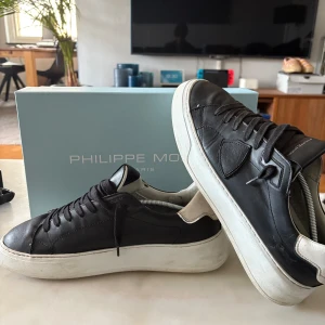 Philippe Model sneakers - Snygga svarta sneakers från Philippe Model med vit platt sula och kontrasterande vit häl. Skorna är i skinn och har klassisk snörning samt rund tå. Perfekta för dig som gillar stilrena och trendiga sneakers med premiumkänsla.