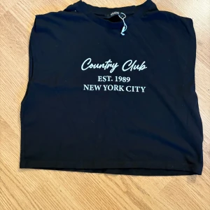 Svart croppad topp från XSIDE - Svart croppad linnetopp från XSIDE med vit text 'Country Club EST. 1989 NEW YORK CITY' framtill. Toppen är ärmlös och har en enkel, rak passform. Perfekt för en chill och avslappnad stil.