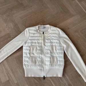 Vit cardigan från Moncler - Säljer en snygg vit kofta från Moncler med quiltad front och två fickor. Koftan har långärmade stickade ärmar och ribbade muddar. Dragkedja framtill och klassisk Moncler-logga. Perfekt för lager-på-lager under kyliga dagar.