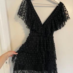 Svart blommig volangklänning Sandro - Supersnygg svart kortklänning från Sandro Paris med blommigt mönster i mesh. Klänningen har v-ringning, volanger och omlottkänsla upptill samt korta, luftiga ärmar. Perfekt för dig som gillar att sticka ut med en cool look.