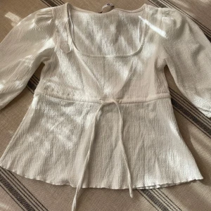 Vit blus med knyt från 157 - Superfin vit blus från 157 med fyrkantig ringning och trekvartsärmar. Toppen har en lätt strukturerad yta och knytband i midjan som ger en snygg siluett. Perfekt att styla med jeans eller kjol för en fräsch look.