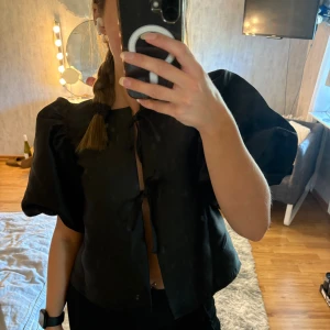 Svart blus med puffärm från Gina Tricot - Supersnygg svart blus från Gina Tricot med stora puffärmar och två knytningar framtill. Blusen är croppad och har en lätt glansig finish som ger en lyxig känsla. Perfekt för dig som vill sticka ut med coola detaljer och volym i ärmarna. Använd två gånger!