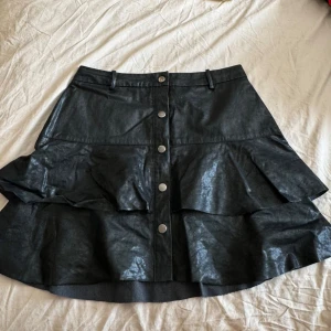 Svart volangkjol i skinnimitation Zara - Supersnygg svart kortkjol från Zara i skinnimitation med volanger och knappar framtill. Kjolen har bälteshällor och en cool, lite glansig finish. Perfekt för dig som vill sticka ut med en edgy vibe. Tyvärr för liten för mig