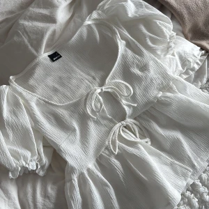 Vit blus med knyt från Gina Tricot - Supersöt vit blus från Gina Tricot med puffiga korta ärmar och två knyt framtill. Blusen har en lätt rynkad struktur och volang nedtill, vilket ger en romantisk vibe. Perfekt för dig som gillar feminina detaljer och en luftig känsla.