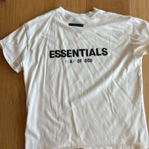 Vit Essentials Fear of God t-shirt - Säljer en clean vit t-shirt från Fear of God Essentials med svart tryck på bröstet. Klassisk rund hals och korta ärmar. T-shirten är gjord i mjuk bomull och har en relaxed passform som är perfekt för en chill look.skriv om ni har frågor 