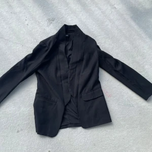 Svart kavaj från SHEIN - Stilren svart kavaj från SHEIN med klassisk krage och två fickor framtill. Kavajen har en enkel, rak passform och är tillverkad i ett mjukt syntetmaterial. Perfekt för dig som gillar en clean look och vill ha något som funkar till många olika outfits.
