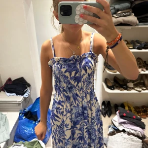 Jättesöt sommarklänning - Supersöt klänning med blått blommönster på vit botten och smockad överdel. Axelbandslös look med tunna axelband och luftig kjol. Perfekt för varma dagar och ger en fräsch vibe. Passar dig som gillar romantisk stil och vill sticka ut med ett färgglatt mönster.