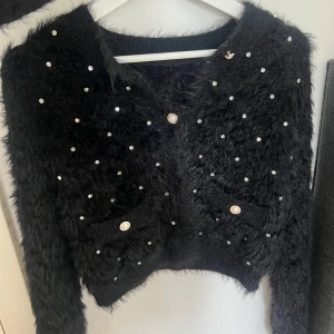 Svart fluffig kofta med pärldetaljer - Supermjuk och fluffig svart kofta med långa ärmar, dekorerad med pärlliknande knappar och glittrande paljetter över hela framsidan. Två små fickor framtill och croppad passform. Perfekt för dig som vill sticka ut med en cozy men glam look.