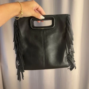 Svart handväska med fransar från Maje - Säljer min Maje väska i skinn i den lite större storleken (inte mini alltså). Väskan har en avtagbar axelrem och kommer med dustbag.