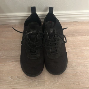 Svarta sneakers från BOSS - Svarta sneakers från BOSS med stickad ovandel och snygga detaljer i syntet. Skorna har snörning, rund tå och platt sula. Perfekta för dig som gillar en stilren och sportig look. BOSS-logga syns både på plösen och hälen.