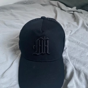 Svart truckerkeps med broderad logga - Snygg svart truckerkeps med broderad logga framtill i svart. Kepsen har baksida för extra ventilation och justerbar rem bak. Perfekt för en clean och stilren look.