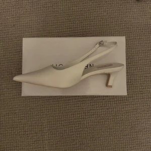 Vita pumps med spetsig tå från Nelly - Stilrena vita pumps från Nelly med spetsig tå och smal rem runt hälen. Skon har en låg, unik klack och är tillverkad i syntetmaterial. Perfekt för dig som vill ha en clean och modern look med en twist.