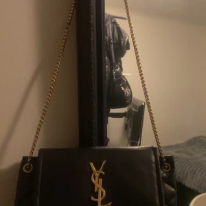 Svart YSL väska - Snygg svart axelväska med guldfärgad kedja och logotypdetalj på framsidan. Väskan har en quiltad design som ger den en lyxig känsla. Perfekt för att lyfta vilken outfit som helst.