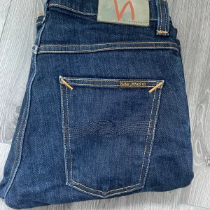 Mörkblå jeans från Nudie Jeans  - Snygga mörkblå jeans från Nudie Jeans  med klassisk femficksdesign och kontrastsömmar. Jeansen har raka ben och subtila detaljer på bakfickorna. Perfekta för dig som gillar stilrena och tidlösa jeans i kraftigt denimtyg. Storlek 30/32, grim tim= slim, riktigt snygga! Hör av dig vid fundering!