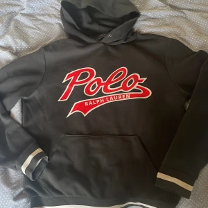 Svart hoodie Polo Ralph Lauren - Svart hoodie från Polo Ralph Lauren med stor röd och vit logga framtill. Klassisk känguruficka, huva och ribbade muddar med vita ränder. Perfekt för en chill och sportig look. Materialet är mjuk bomullsmix som känns skön mot huden. Har haft ett minimalt hål vid fickan som man kan se på bilden men tack vare bra syning är det svårt att se om man inte är nära annars är den jätte bra kond 8/10