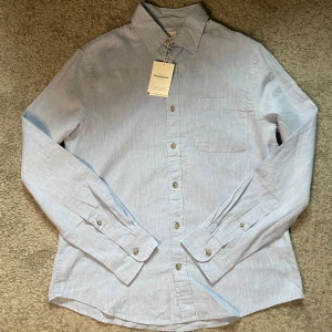 Ljusblå linneskjorta från Dressmann - Snygg ljusblå skjorta från Dressmann i linneblandning. Klassisk krage, bröstficka och knäppning framtill. Perfekt för dig som gillar en stilren och fräsch look. Långärmad modell med normal passform.