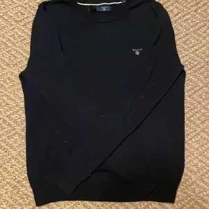 Svart långärmad tröja från GANT - Svart långärmad tröja från GANT med diskret logga broderad på bröstet. Klassisk rund halsringning med ribbade muddar. Perfekt för dig som gillar stilrena och enkla plagg med en snygg passform.