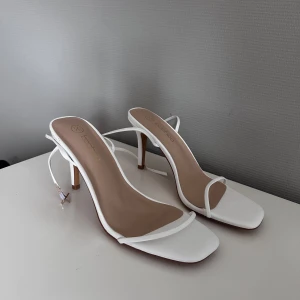 Vita klackskor från Boohoo - Stilrena vita klackskor från Boohoo med smala remmar och fyrkantig tå. Skorna har hög klack och tunna snören som knyts runt vristen för en trendig look. Perfekta för dig som gillar minimalistisk och modern stil.