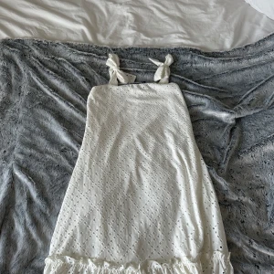 Vit kort klänning från Vero Moda - Supersöt vit klänning från Vero Moda i storlek XS. Klänningen har knytband på axlarna, broderade hålmönster och en volangkant längst ner. Perfekt för dig som gillar romantisk och feminin stil. Materialet känns lätt och luftigt, perfekt för varma dagar. Aldrig använd, Köpte för att använda på avslutning men blev aldrig så, så den är aldrig använd😊