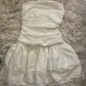 Vit bandou kort volang klänning - Säljer en söt vit axelbandslös klänning med broderade hålmönster och spetsdetaljer längs nederkanten. Klänningen har en volangig kjol och är perfekt för varma dagar. Den är kort och har en romantisk vibe som passar dig som gillar ljusa och fräscha looks. Aldrig använd 