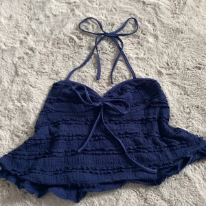 Mörkblå croppad linnetopp med knyt - Supersöt mörkblå croppad linnetopp med volangdetaljer och knyt både i nacken och framtill. Toppen har en lätt strukturerad yta och är perfekt för varma dagar. Sitter snyggt med sin ärmlösa design och ger en chill vibe. Har använt två gånger.