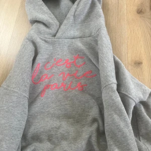 Grå hoodie med rosa text från Uniq Young - Säljer en grå hoodie från Uniq Young med rosa texten 'c'est la vie paris' på bröstet. Tröjan har huva och är långärmad, perfekt för en avslappnad stil. Mjuk och skön bomullsblandning.💖