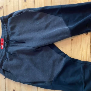 Nike teech mjukisbyxor i svart och grå - Nike mjukisbyxor i svart och mörkgrå med snygg tvåfärgad design. Byxorna har dragsko i midjan, dragkedjeficka och är gjorda i mjukt bomullsmaterial. Perfekta för chill eller träning, med klassisk Nike-logga på benet.