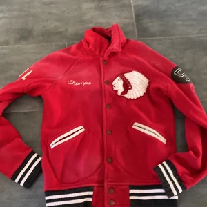 Röd bomberjacka Polo Ralph Lauren S - Röd bomberjacka från Polo Ralph Lauren med broderade detaljer, stor logga på ryggen och coolt indiantema på bröstet. Jackan har svarta och vita ribbade muddar, knappar framtill och två fickor. Perfekt statement-plagg.