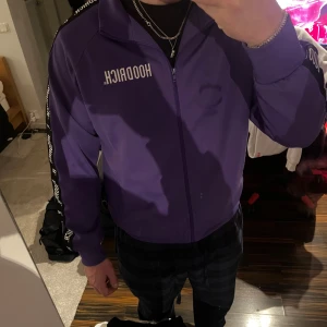 Lila track jacket från Hoodrich - Säljer en lila track jacket från Hoodrich med dragkedja framtill och svarta band med vit logga längs ärmarna. Jackan har hög krage och lång ärm, perfekt för en sportig och trendig look. Passar M/L! 