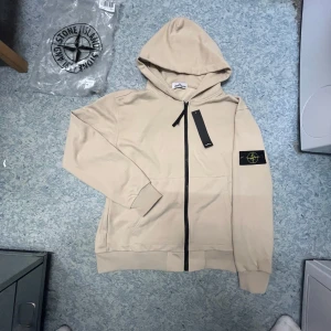 Beige hoodie från Stone Island L - Snygg beige hoodie från Stone Island med klassisk patch på ärmen. Tröjan har huva, hel dragkedja framtill och ribbade muddar. Tillverkad i mjuk bomull, perfekt för en chill och stilren look. Svarta detaljer vid dragkedjan ger extra edge.