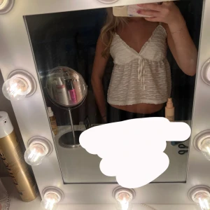 Vit linnetopp med knyt och struktur - Supersöt vit linnetopp med smala axelband och v-ringning. Toppen har en vågig, strukturerad yta och en söt knytning framtill. Lätt och luftig modell som är perfekt för varma dagar. Passar dig som gillar en feminin och trendig stil.