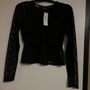 Svart spetsblus från Gina Tricot - Supersnygg svart blus i spets från Gina Tricot. Modellen har långa ärmar, rund halsringning och lätt volang nertill. Toppens genombrutna spets ger en cool och trendig look, perfekt att styla med jeans eller kjol.
