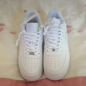 Nike Air Force 1 helvita sneakers - Klassiska Nike Air Force 1 sneakers i helvitt skinn med perforerad tå, vit sula och snörning. Swoosh-logga på sidan och metallbricka på snörningen. Perfekta för en clean och stilren look.