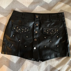 Svart kortkjol med pärlor och nitar - Snygg svart kortkjol i skinnimitation med dekorativa pärlor och nitar på fickorna och framtill. Kjolen har två fickor med lock, knappar längs hela framsidan och bälteshällor. Perfekt för dig som gillar edgy detaljer och vill sticka ut.