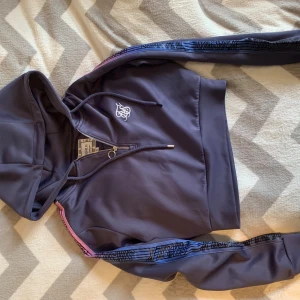 Blå croppad hoodie från SikSilk - Säljer en blå croppad hoodie från SikSilk med dragkedja och snörning i huvan. Tröjan har långa ärmar med blå och rosa detaljer samt logotyp på bröstet. Materialet är mjukt och känns sportigt, perfekt för chill eller träning.